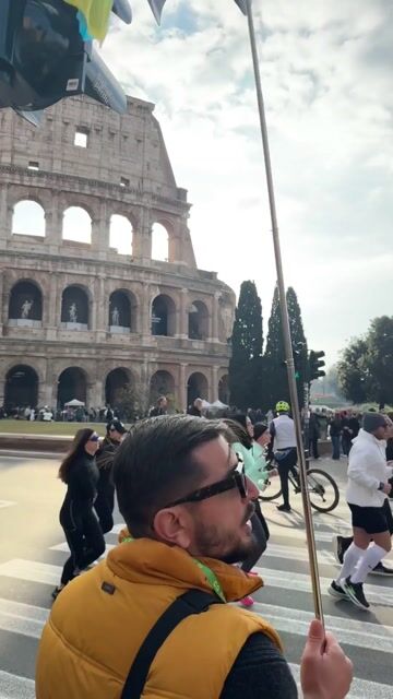 Tourists visiting the Colosseum in Rome, with a blue and yellow flag on a flagpole in the foreground.