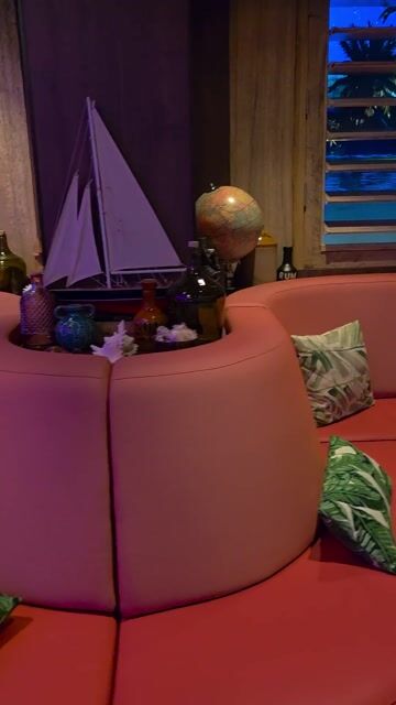 A modern nightlife venue with vibrant purple and green neon lighting, tropical plants, a red curved seating area, bar seating, and people socializing in an upscale lounge environment.