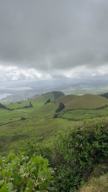 Rolling green hills with scattered trees and lush vegetation in the foreground under a partly cloudy sky.