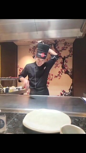 A chef demonstrates a cooking technique at an open kitchen counter with decorative floral wall art in the background.